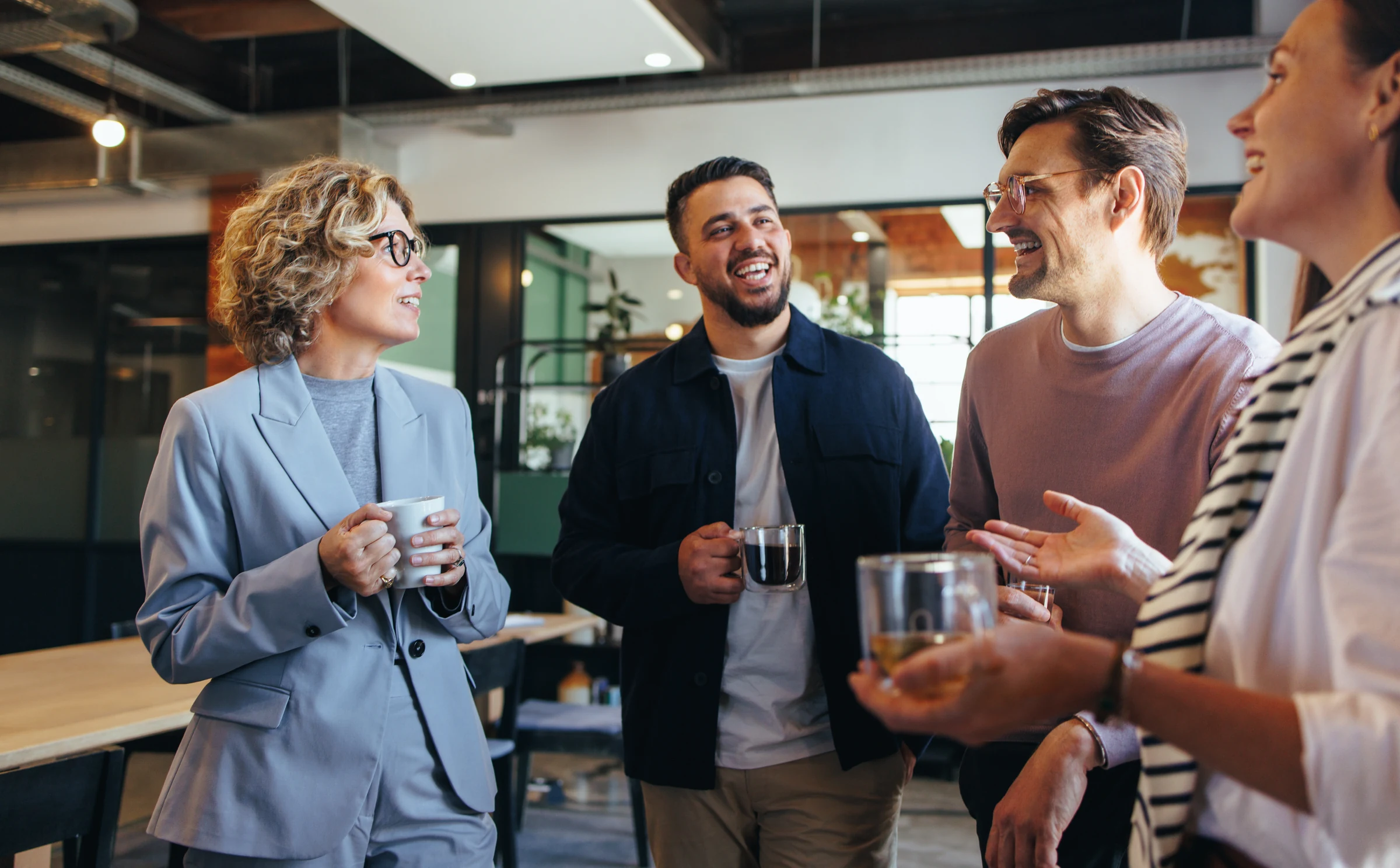 Four colleagues talking and smiling together over coffee in a shared workspace