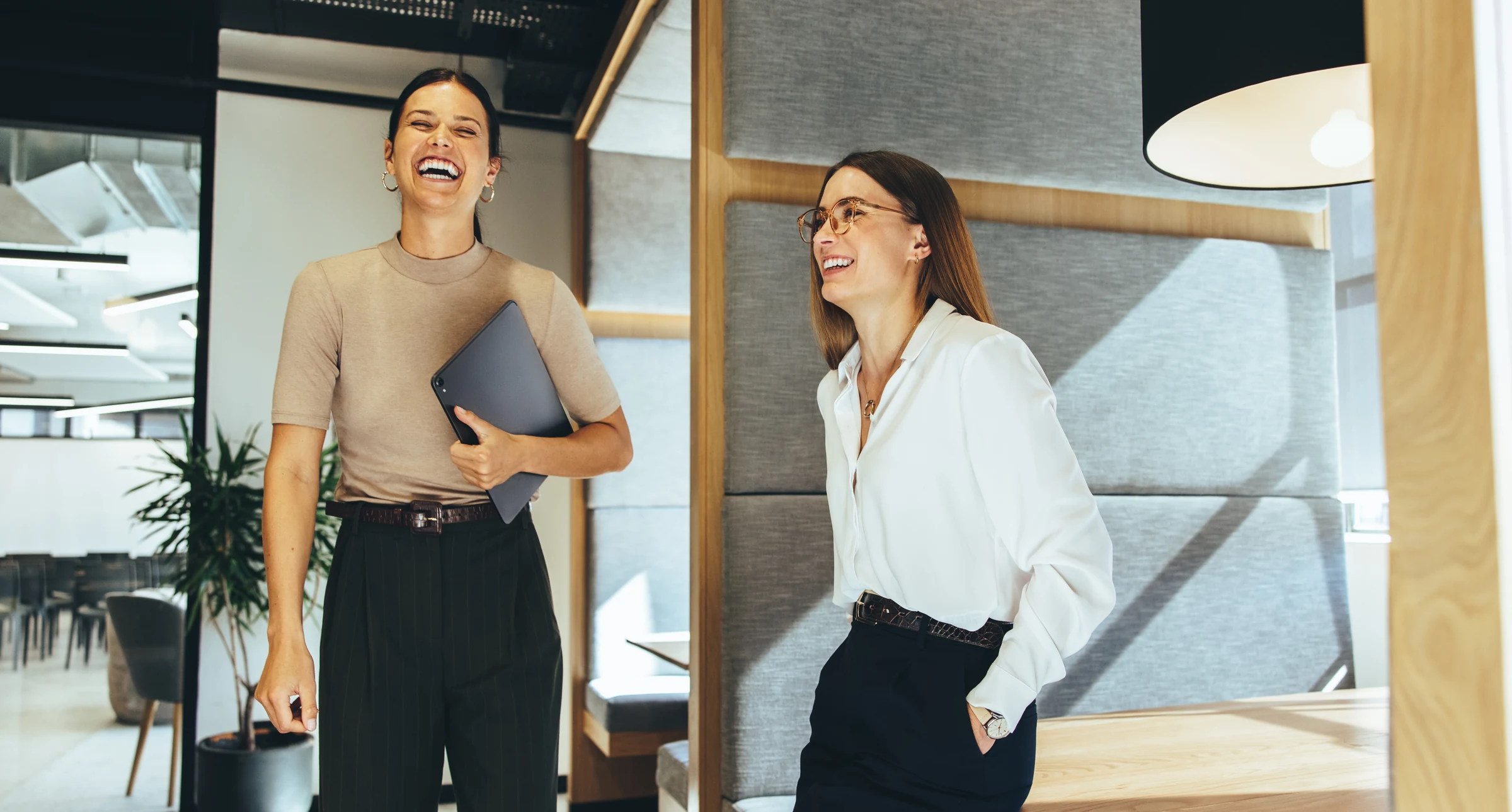 Two colleagues sharing a laugh in a bright workspace, one holding a tablet