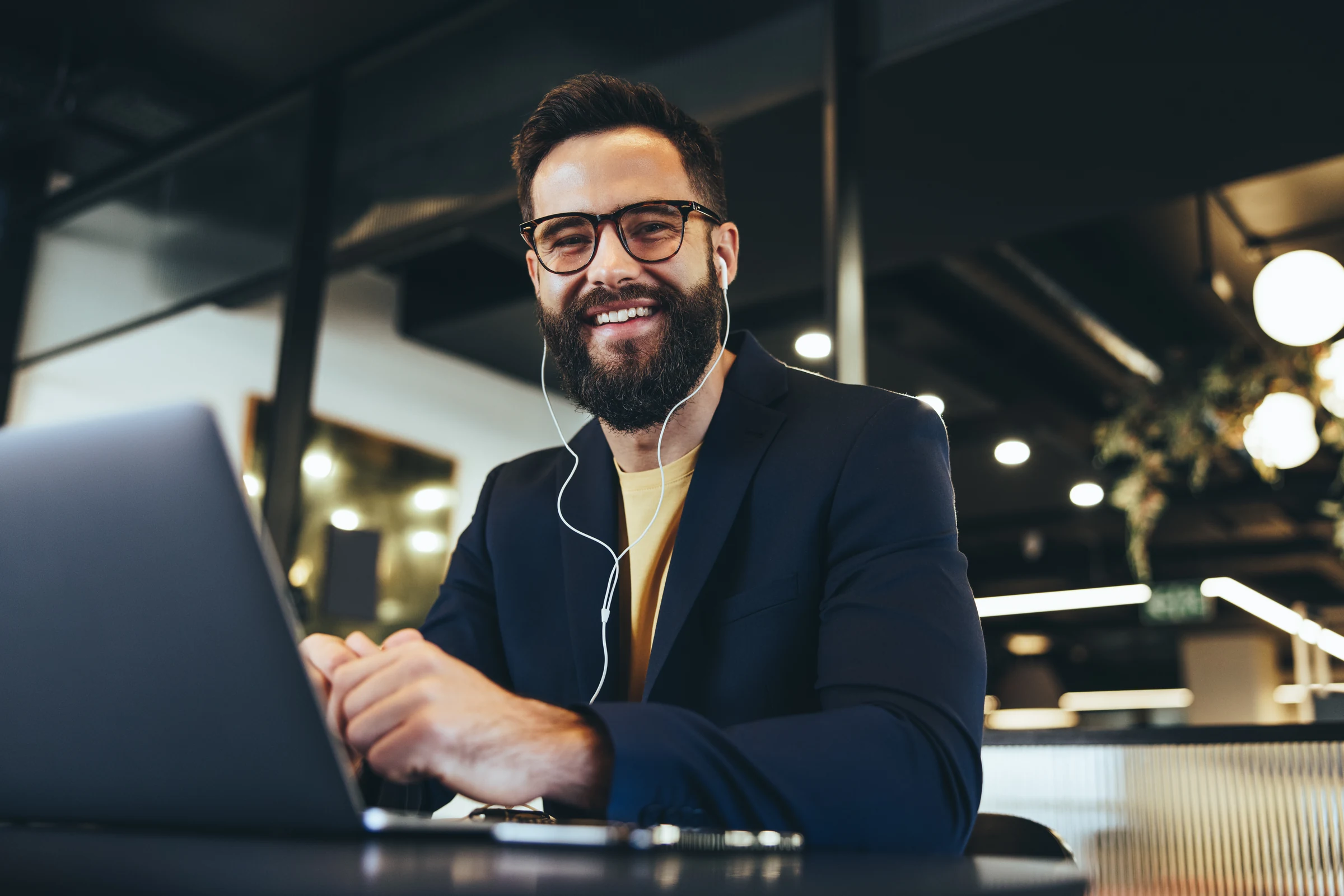 A professional wearing earphones and a navy jacket using a laptop in a warmly lit shared workspace