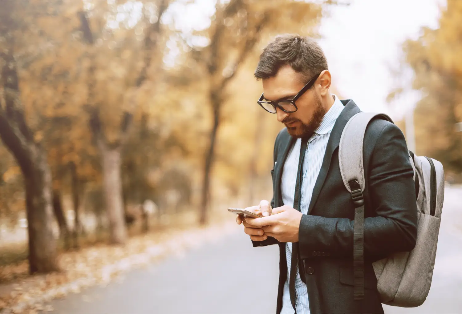 A man with glasses and a backpack using his phone outdoors in autumn