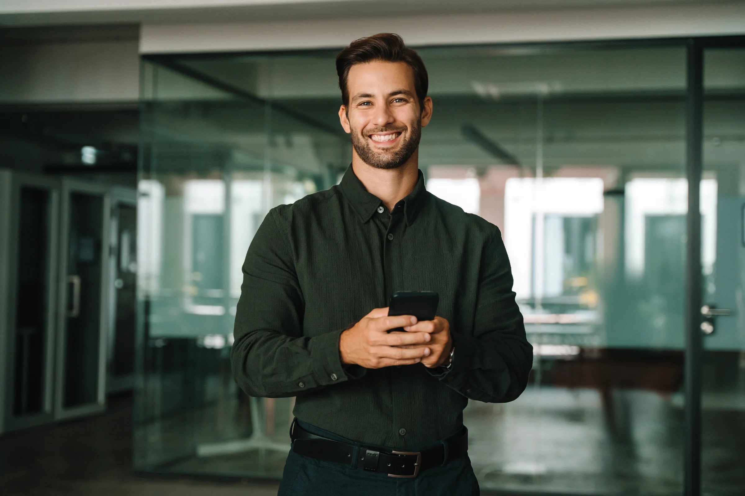 A smiling professional in a dark shirt holding his smartphone in an office