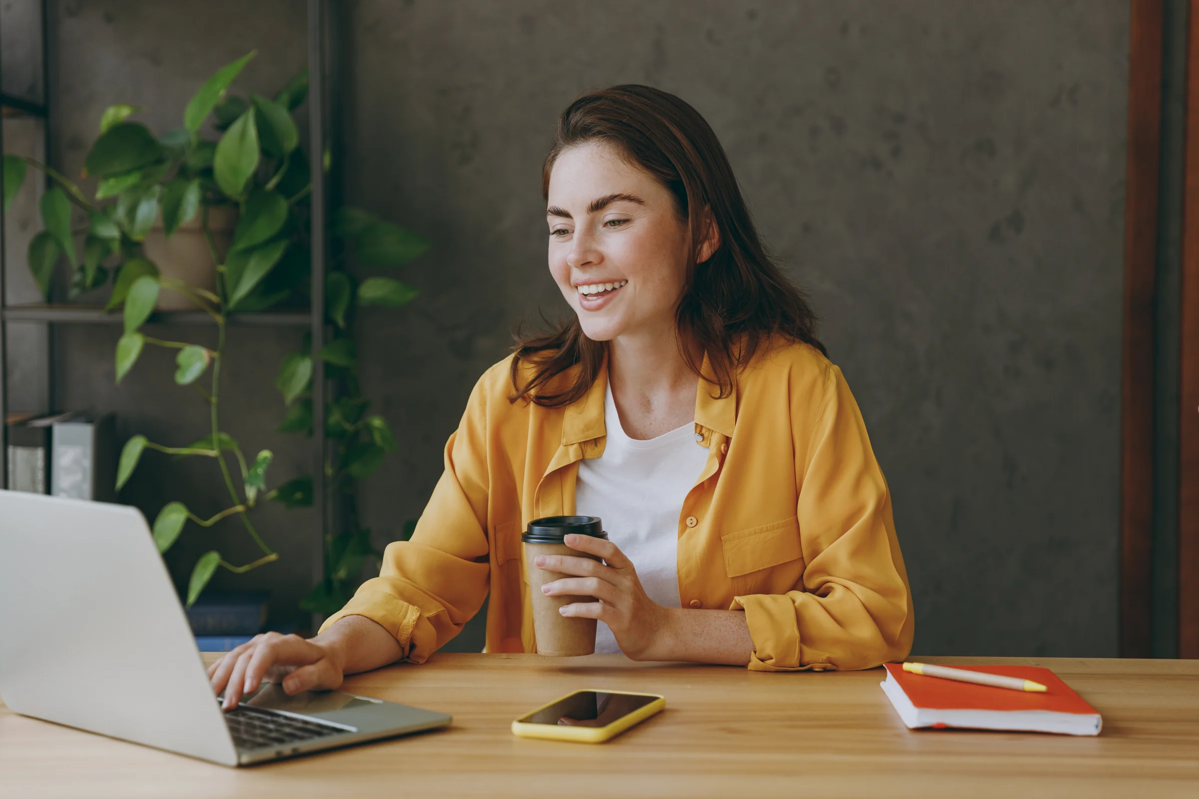 A woman in a yellow shirt working on a laptop with a coffee beside her.