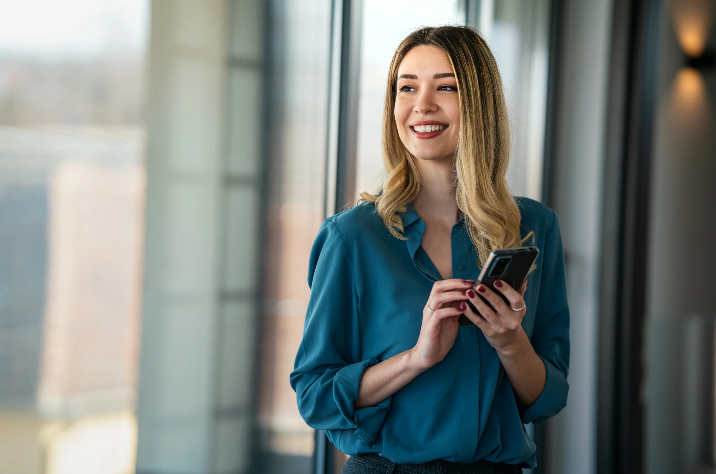 A professional in a teal blouse holding a smartphone beside an office window