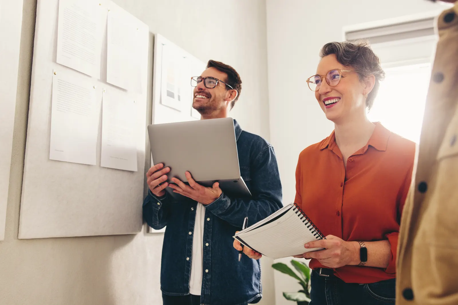 Colleagues with a laptop and notebook beside planning notes on an office wall
