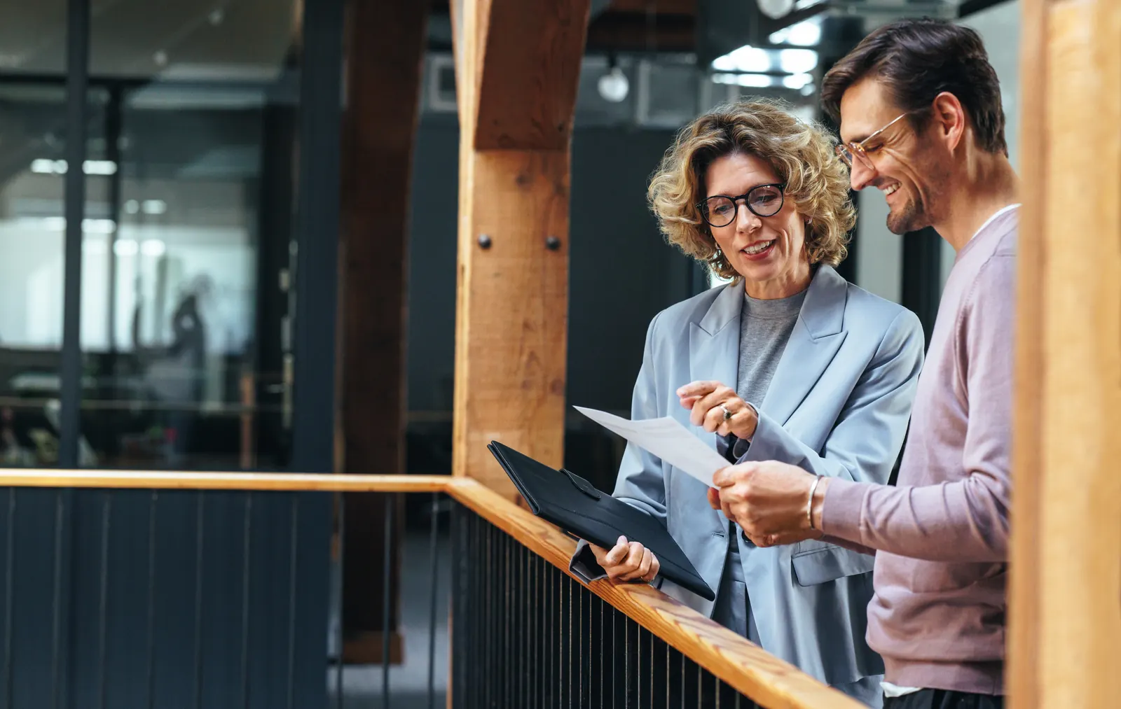 Two colleagues reviewing a tablet and printed notes together beside a wooden office railing