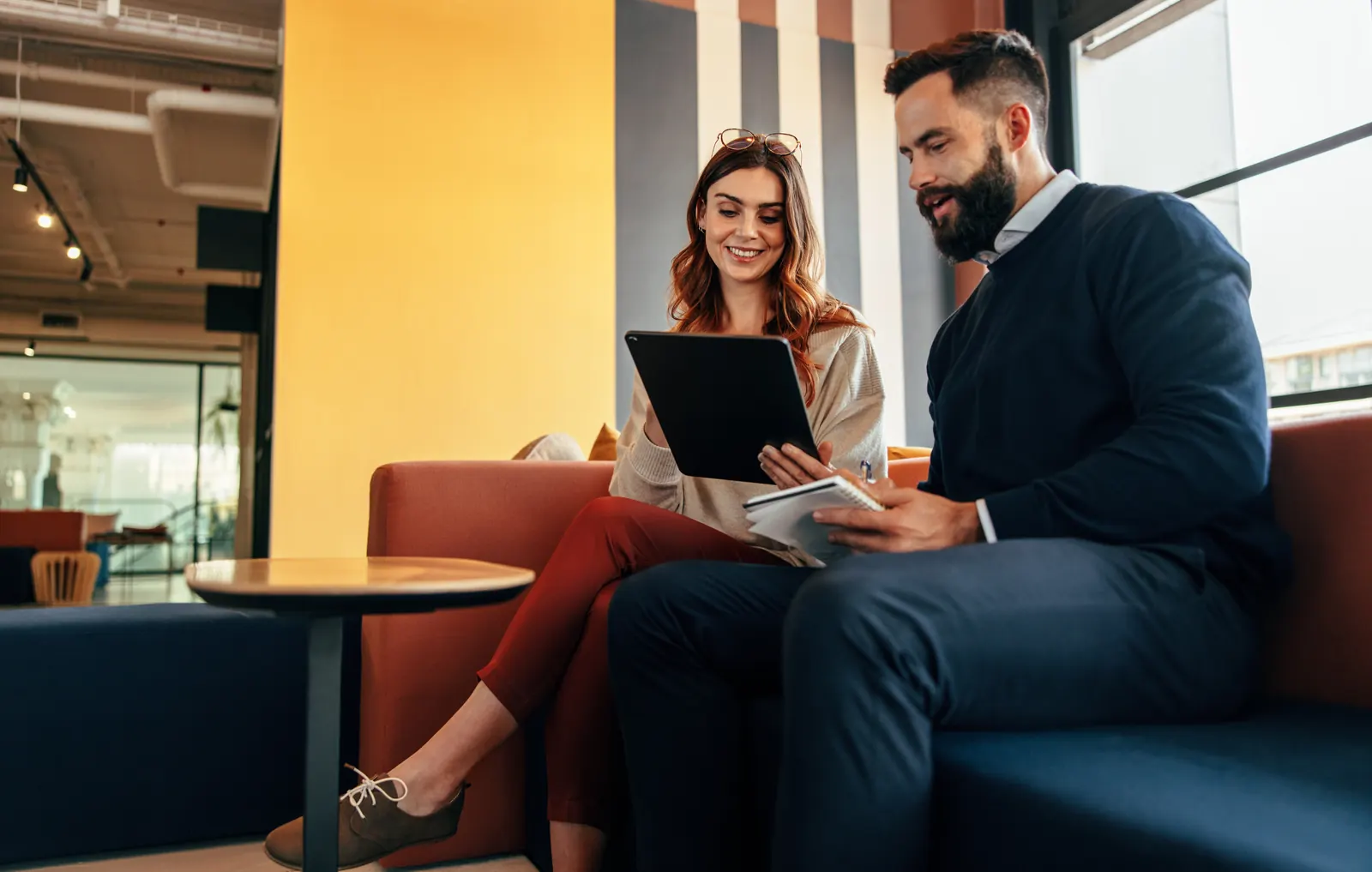 Two colleagues reviewing a tablet and notes in an office lounge.