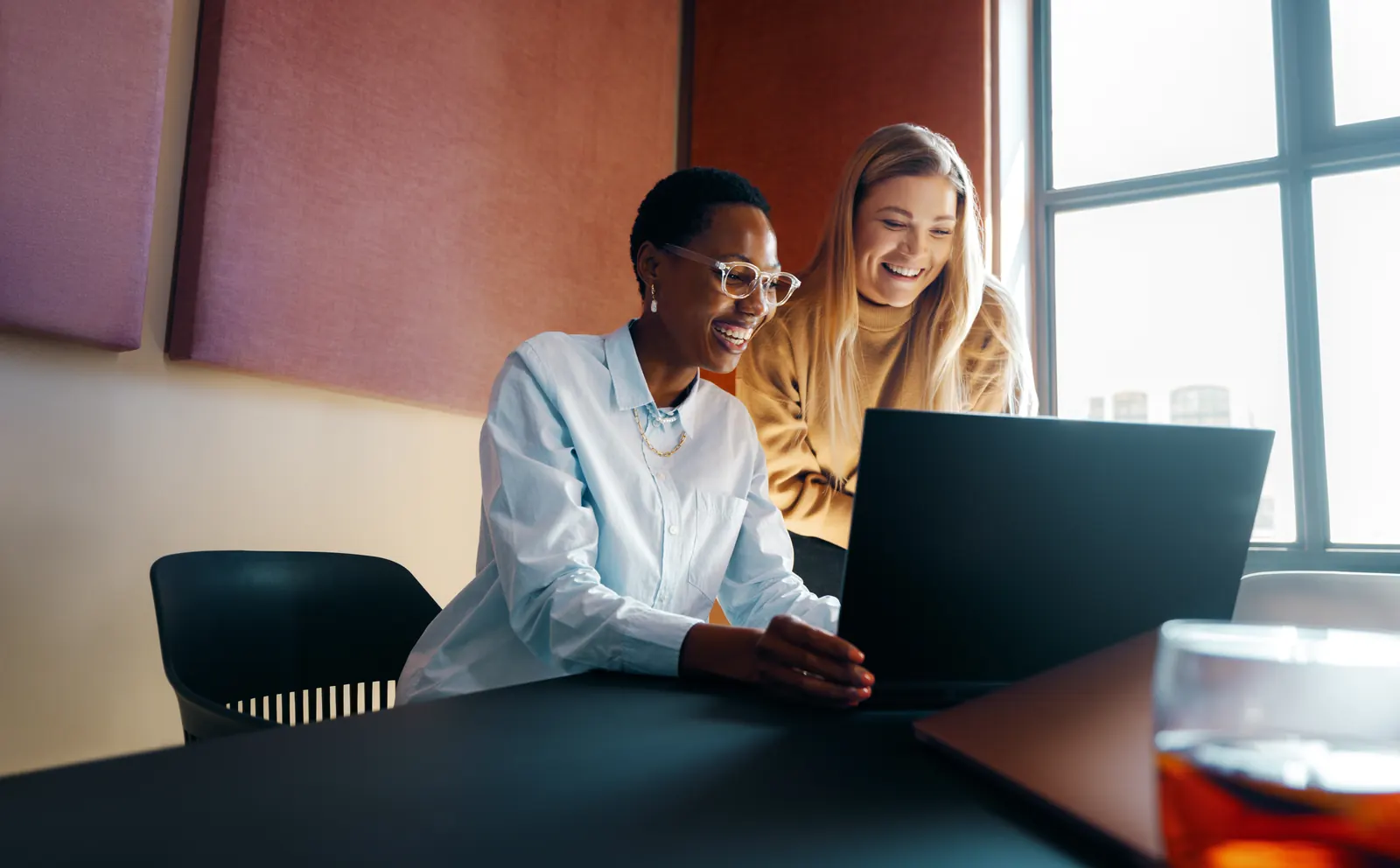 Two colleagues working together on a laptop beside a sunlit office window.