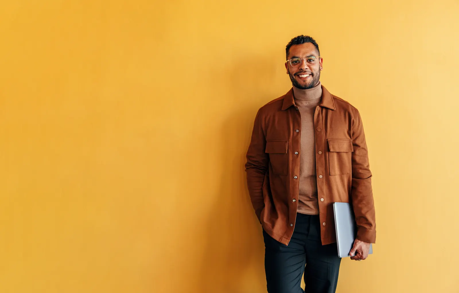A business professional in a burnt-orange jacket holding a closed laptop against a warm background