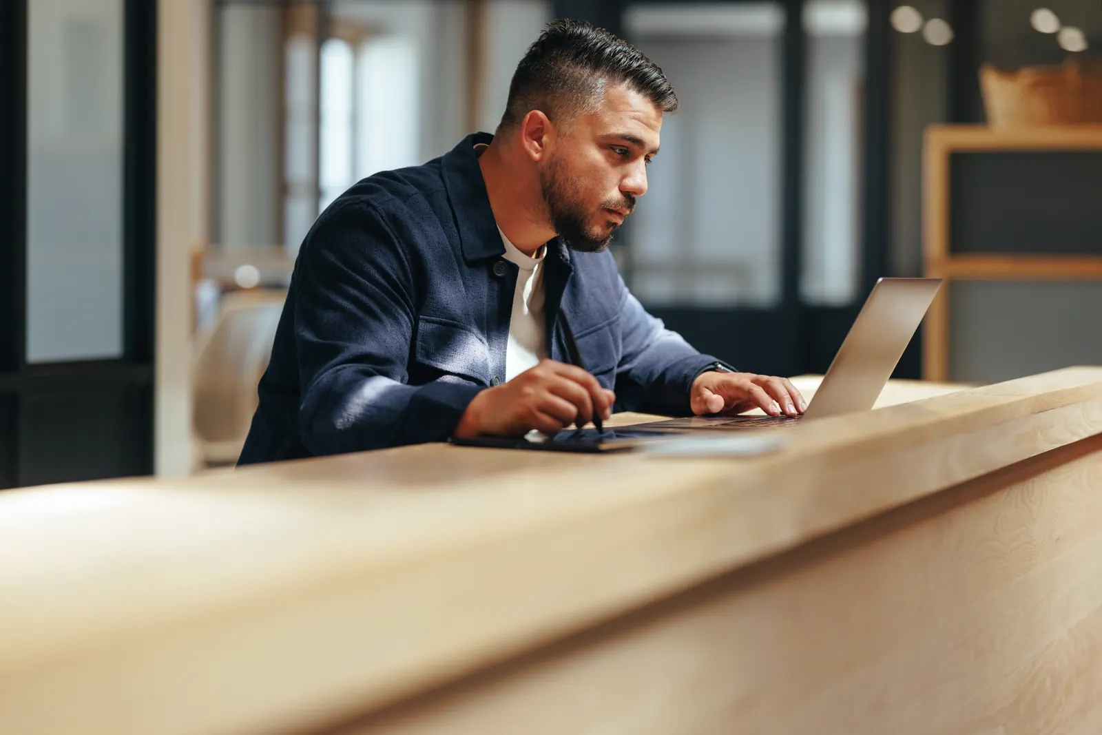 A professional in a navy shirt working between a laptop and tablet at a warm wooden desk