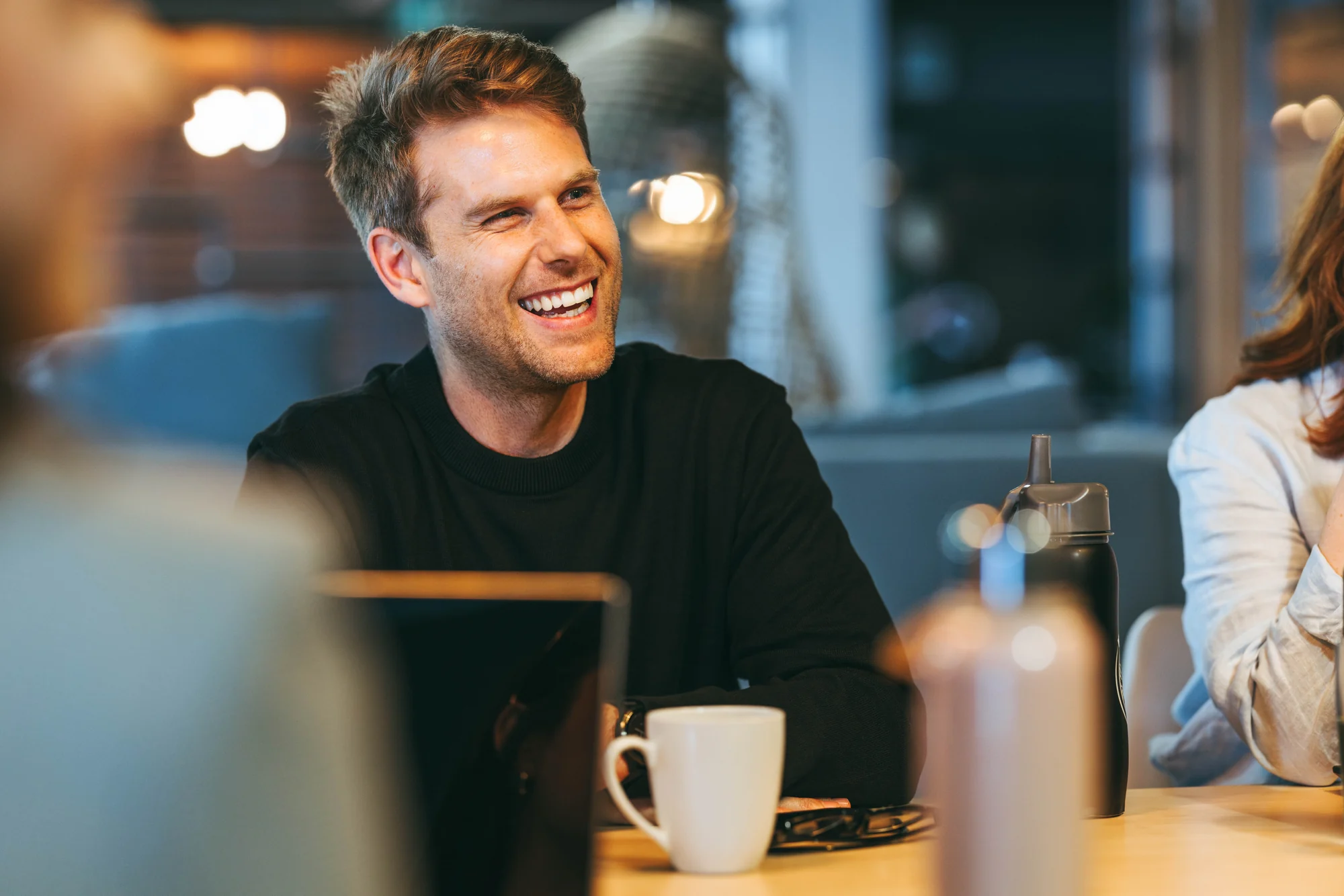 A smiling colleague in conversation at a meeting table with a laptop and coffee in a naturally lit office