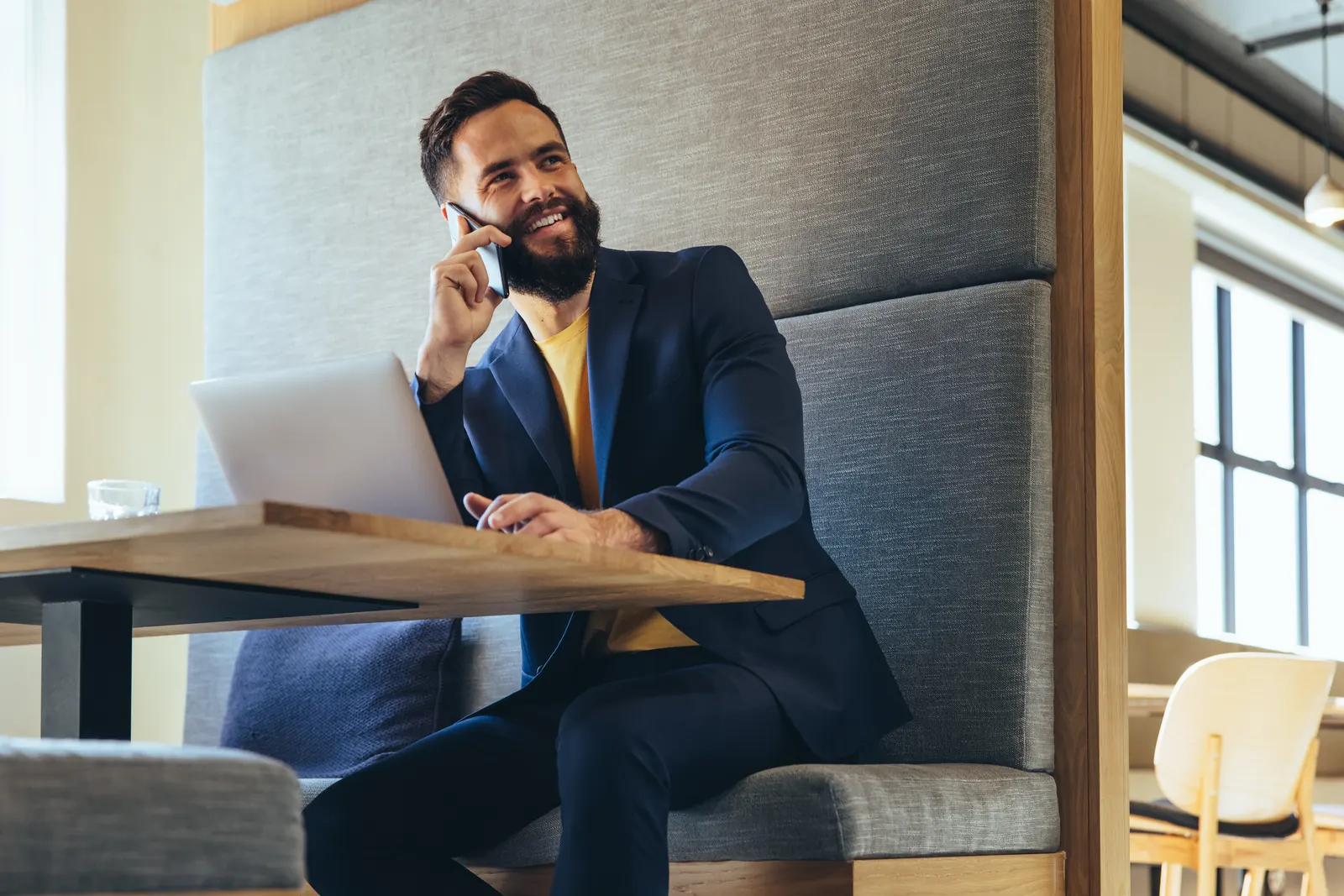 A professional in a navy jacket on a phone call beside his laptop in a quiet office booth.