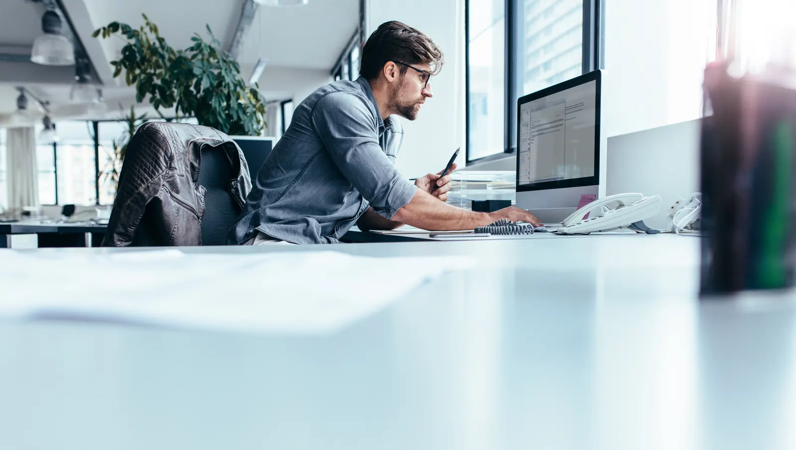 A professional using a desktop computer while holding a phone in a bright office