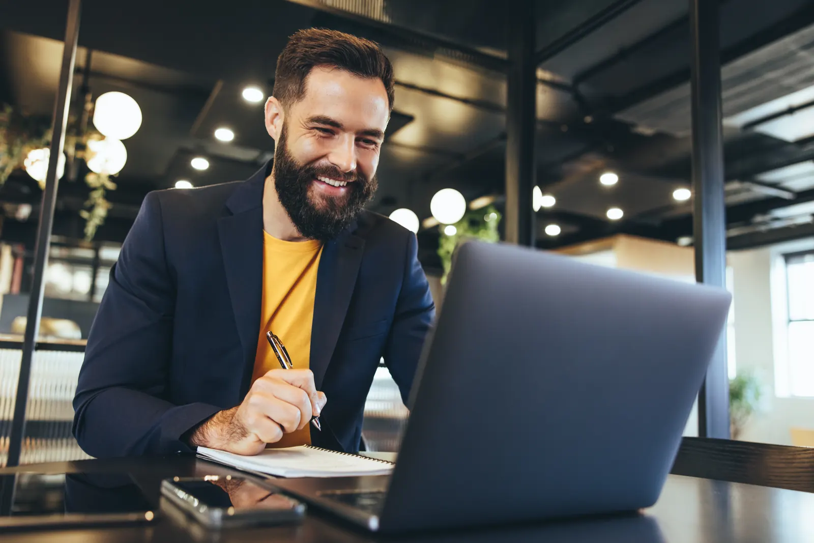 Professional in a navy blazer taking notes beside a laptop in a shared workspace