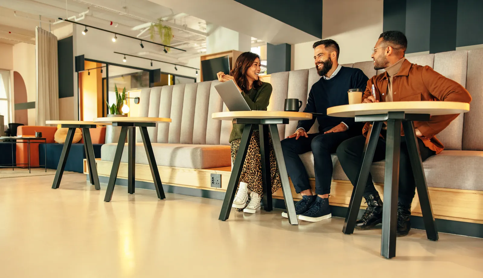 Three colleagues talking at small tables with a laptop in an office lounge