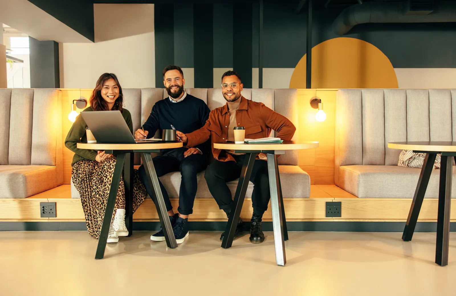 Three colleagues seated at small tables with a laptop in an office lounge