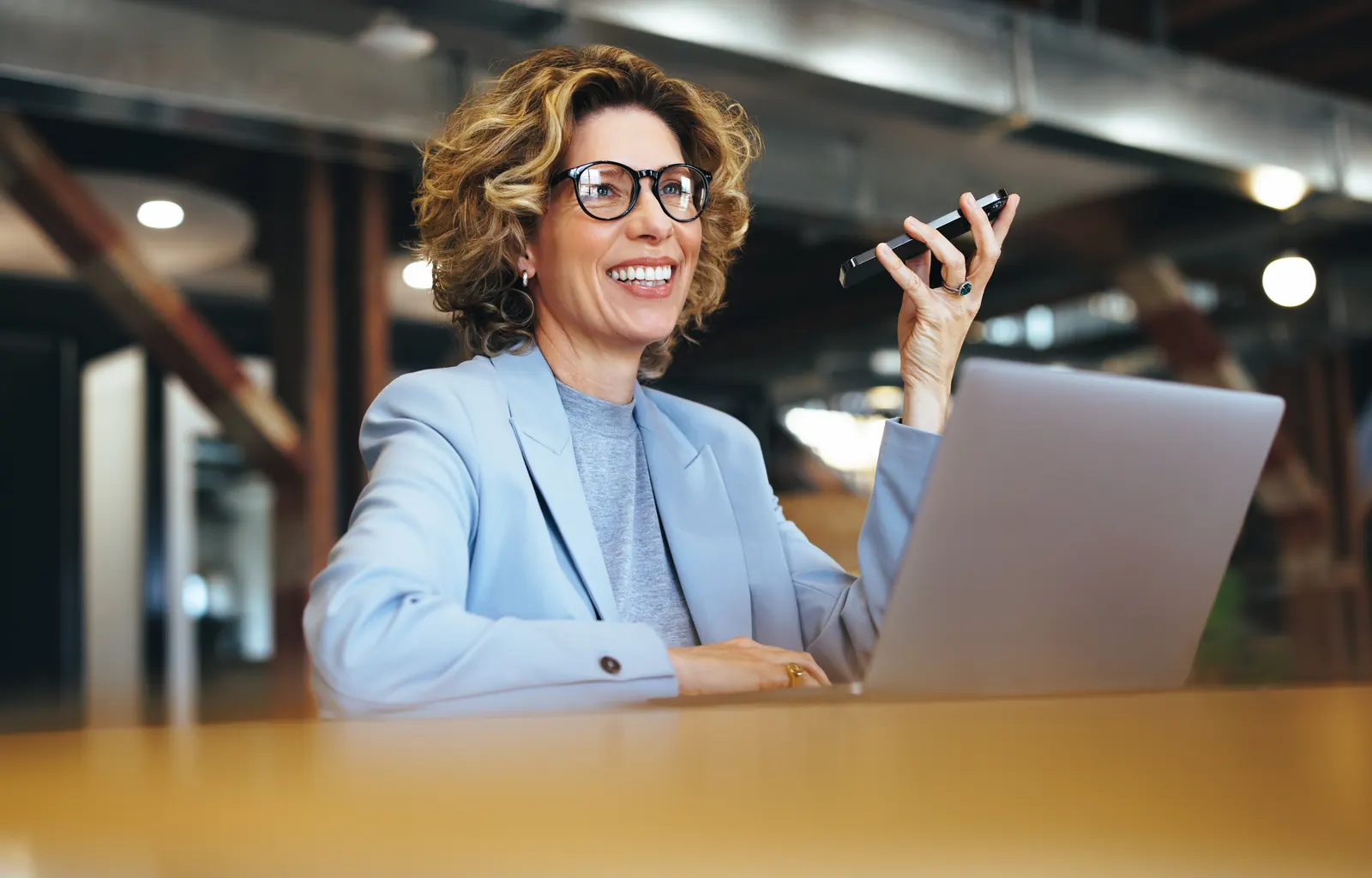 Professional in a blue blazer speaking on a phone beside a laptop in a warm shared workspace