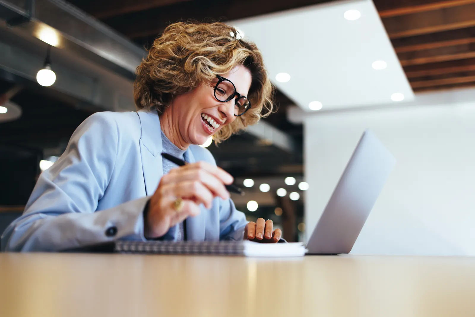 A smiling professional in a blue shirt taking notes beside her laptop